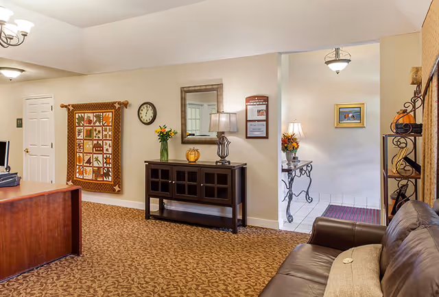 A senior living facility reception/lounge showing a leather sofa, wooden desk, console table with lamp and mirror, wall clock and decorative quilt, and an open tiled entryway.