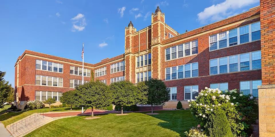 Exterior view of a large, historic brick building with multiple windows and decorative architectural details. The building is surrounded by well-maintained green lawns, trees, and flowering bushes under a clear blue sky.