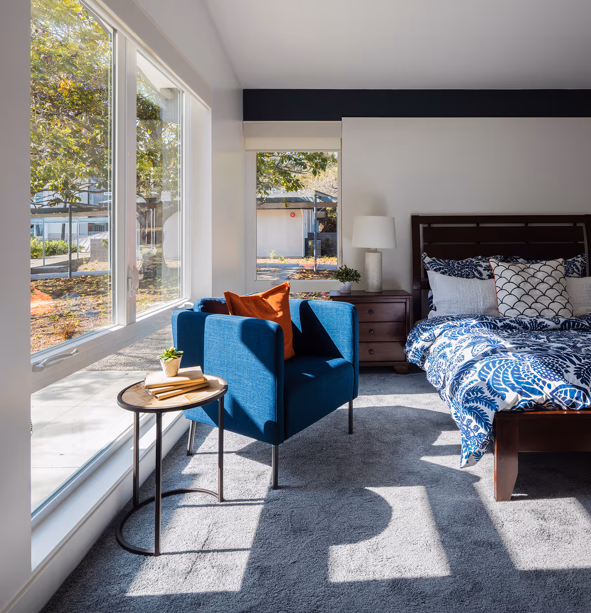 A bright bedroom with a large window letting in natural light. The room features a dark wooden bed with blue and white patterned bedding and multiple pillows. Next to the bed is a wooden nightstand with a white lamp and a small plant. In front of the window, there is a blue armchair with an orange pillow and a small round side table holding a small plant and some books. The floor is carpeted in light gray.