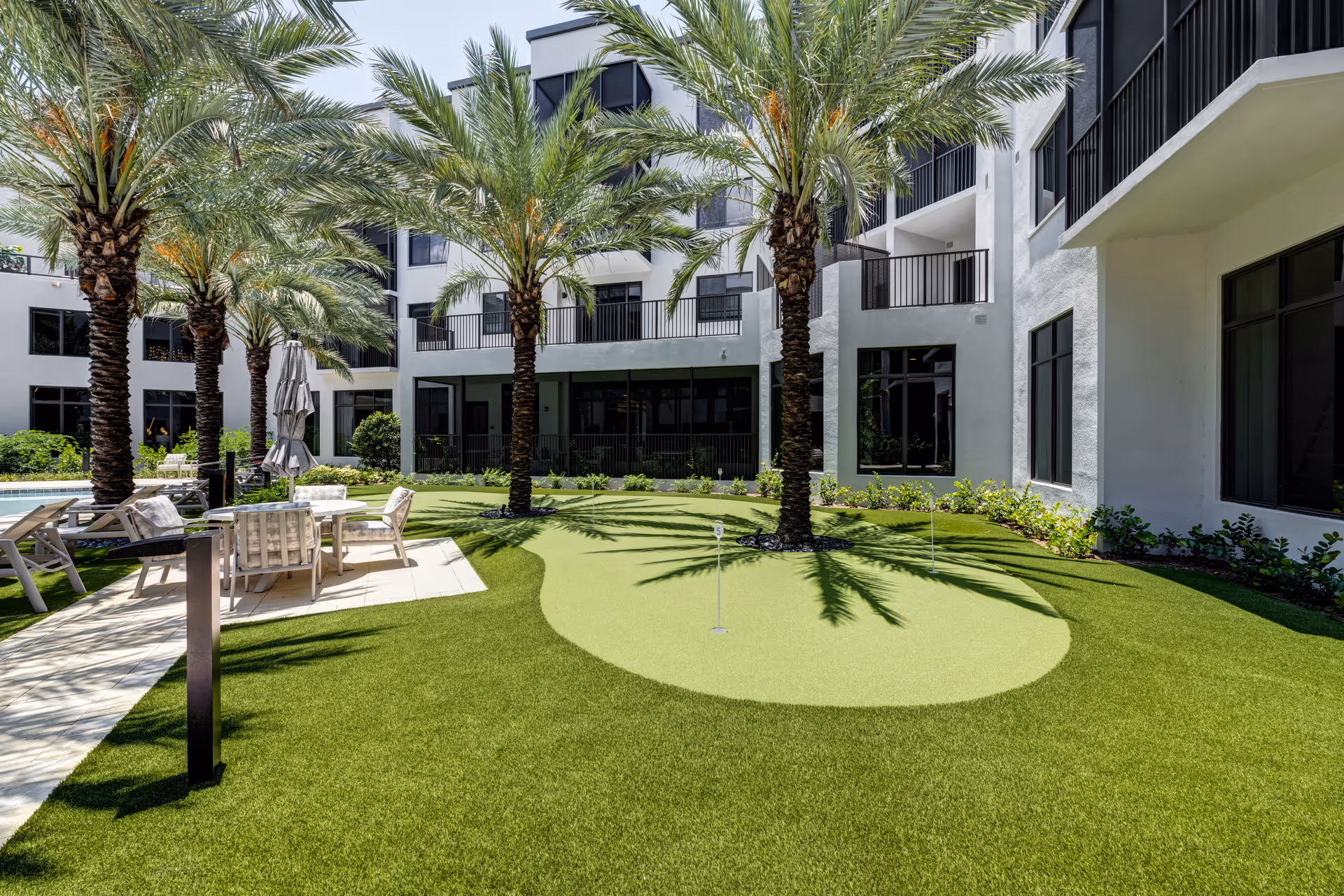 Sunlit courtyard with palm trees, a small putting green, and patio seating surrounded by a multi-story building.