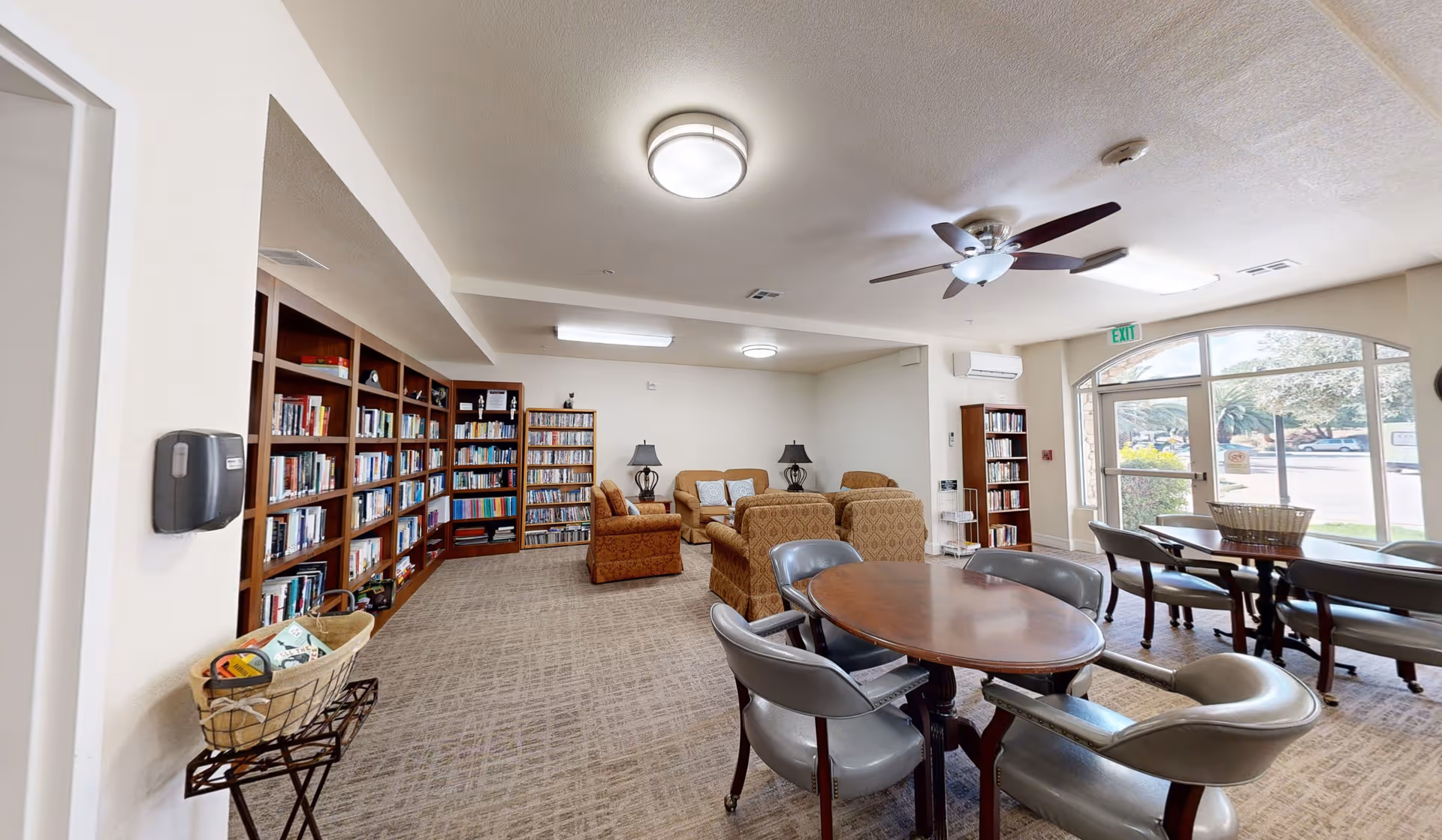 A well-lit common area in a senior living facility featuring bookshelves filled with books along the left wall, several upholstered armchairs and a sofa arranged around a small table with two lamps, and multiple round tables with chairs near large windows letting in natural light.