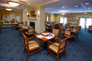 A spacious dining room with multiple wooden tables and cushioned chairs arranged neatly. The room features a blue carpet with a patterned design, yellow walls, and large windows with curtains allowing natural light to enter. There is a fireplace with a painting above it and decorative wall sconces on the walls.