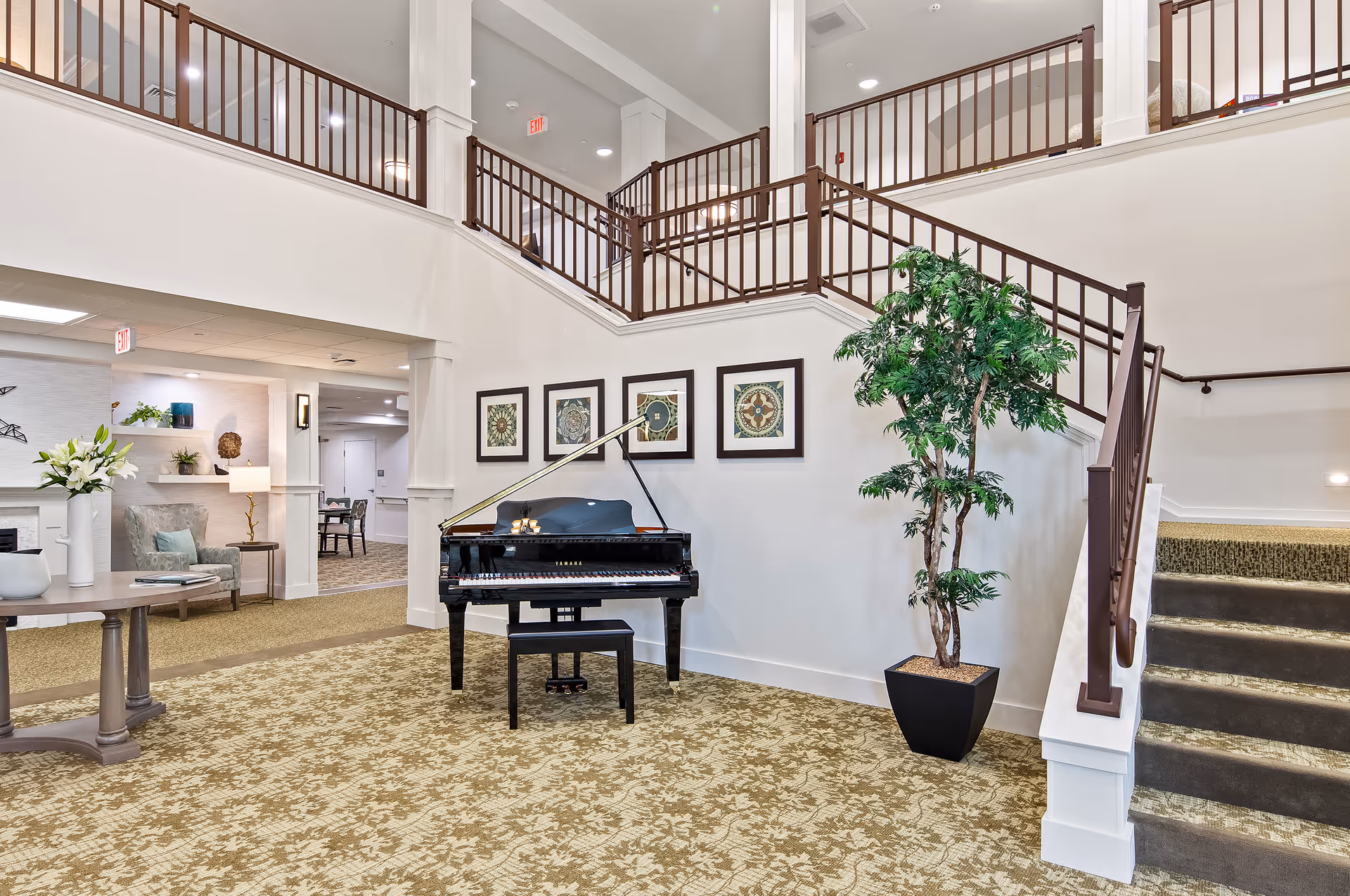 Bright communal lobby with a black grand piano, staircase, framed art, and a potted plant.