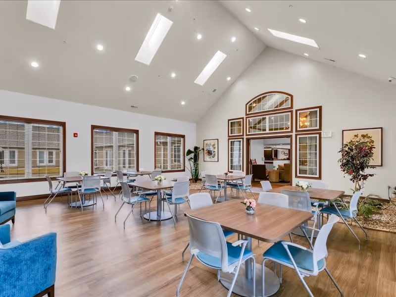 A bright and spacious dining area with multiple square wooden tables and blue and white chairs arranged neatly. The room has large windows with wooden frames, allowing natural light to fill the space. The ceiling is vaulted with recessed lighting and skylights. There are plants and framed artwork on the walls, and an open doorway leads to a cozy living area with sofas.