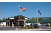 Front entrance of a single-story care facility with two flagpoles, a covered drop-off, parking area, and mountains in the background.