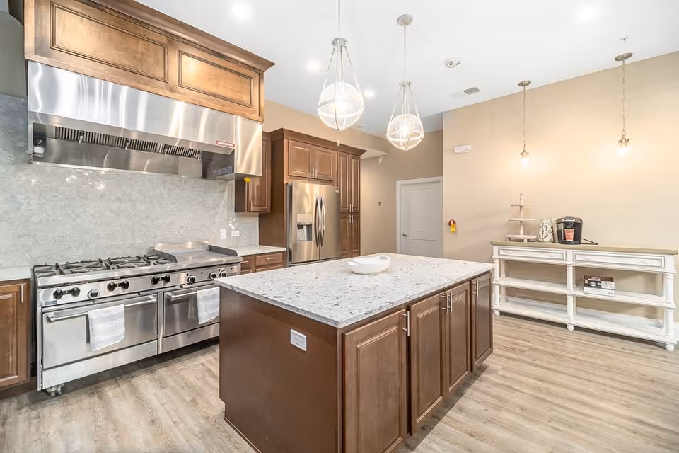 A modern kitchen with a large central island featuring a white marble countertop and dark wooden cabinets. The kitchen includes a stainless steel commercial-grade stove with two ovens and a large stainless steel range hood above it. There is a stainless steel refrigerator and additional wooden cabinetry along the wall. Three pendant lights hang from the ceiling, and a white sideboard with a coffee maker and tiered tray is against the far wall. The floor is light wood, and the walls are painted a light beige color.