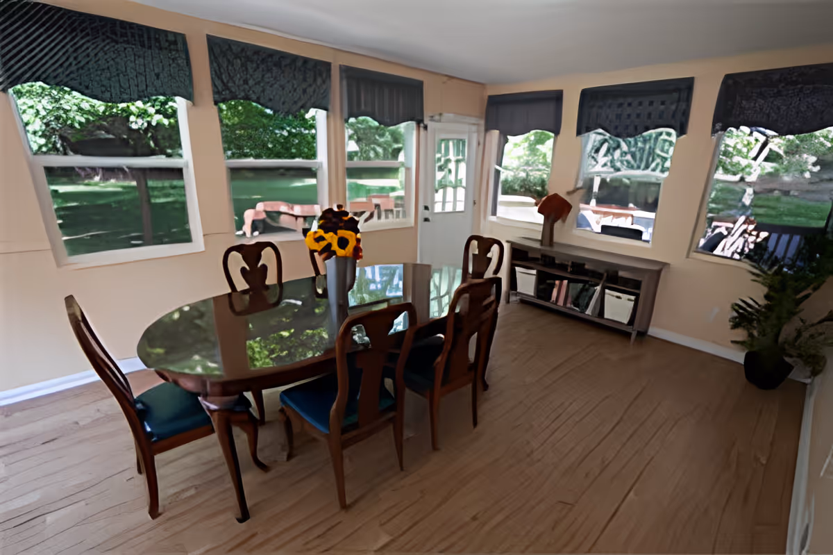 A dining room with a wooden oval table surrounded by six chairs with blue cushions. A vase with sunflowers is placed on the table. The room has multiple windows with dark blue valances, allowing natural light to enter and providing a view of a green outdoor area. There is a sideboard with decorative items and a potted plant in the corner.