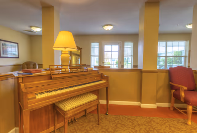 A cozy interior space featuring a wooden upright piano with a cushioned bench, a table lamp on top of the piano, and a red upholstered armchair to the right. The room has beige walls with white trim and large windows letting in natural light.