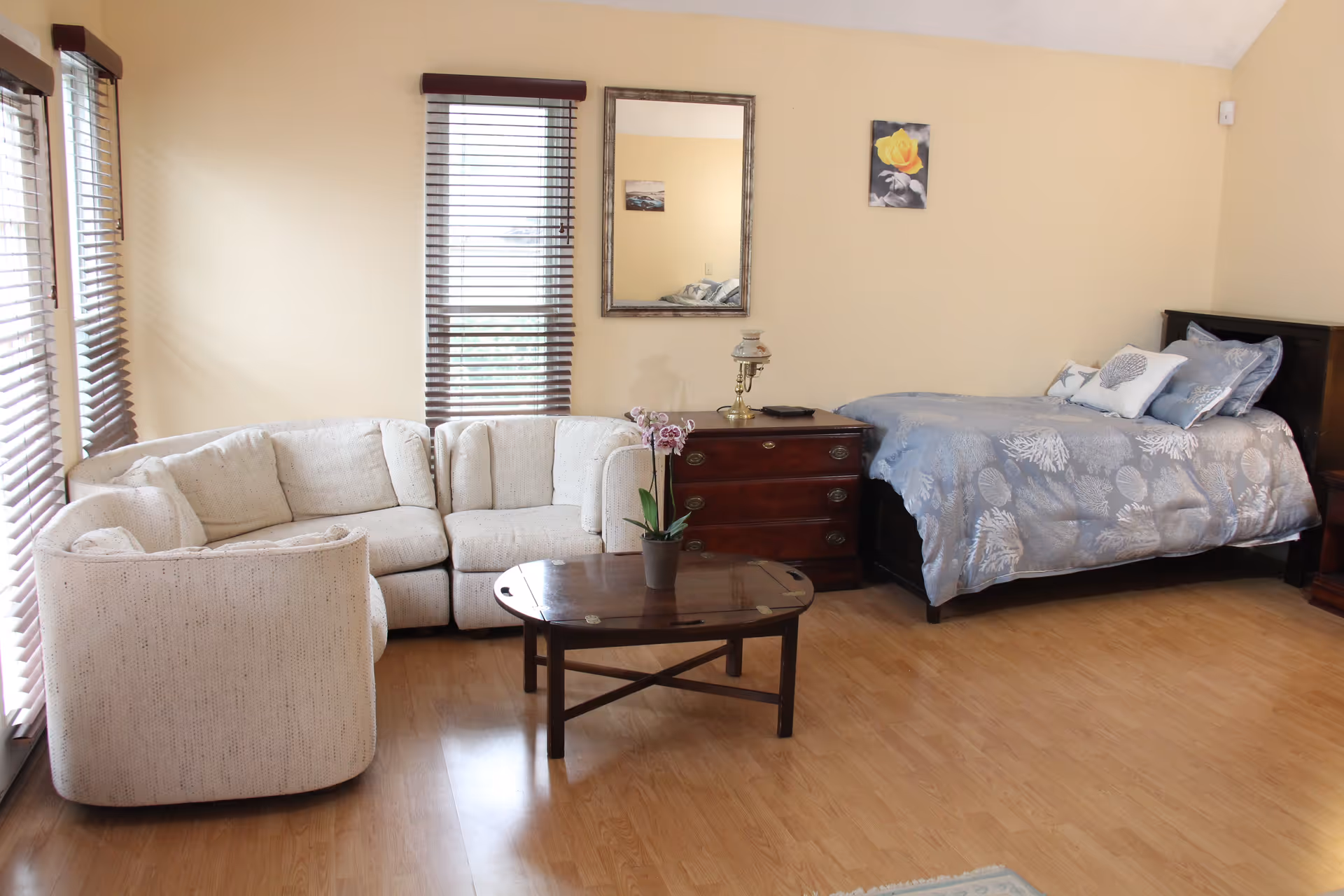 A cozy room with a light beige sectional sofa, a wooden coffee table with a potted orchid, a wooden dresser with a vintage lamp, and a single bed with blue and white patterned bedding. The walls are light yellow with two windows covered by brown blinds, a mirror, and two small paintings.