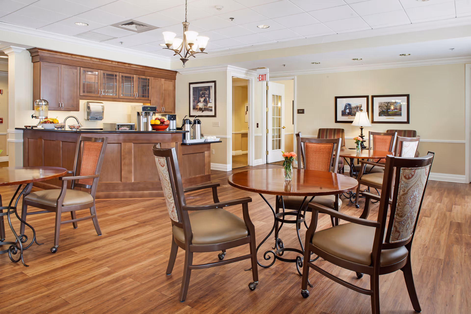 A bright and clean dining area in a senior living facility with wooden tables and chairs arranged neatly. There is a wooden counter with a coffee machine, fruit bowls, and beverage dispensers. The room has wood flooring, framed pictures on the walls, and a chandelier hanging from the ceiling.