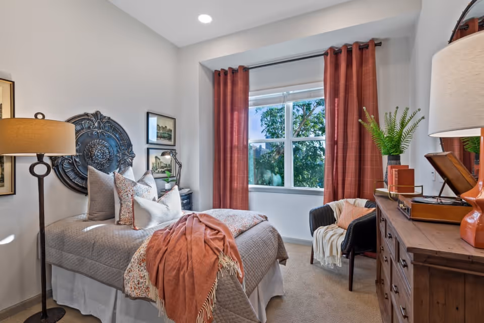 A cozy bedroom with a neatly made bed featuring multiple pillows and a coral throw blanket. The room has a large window with coral-colored curtains, a black chair with a throw blanket and pillow, a wooden dresser with a lamp and decorative items, and a floor lamp beside the bed. The walls are decorated with framed artwork and a large decorative metal piece above the bed.
