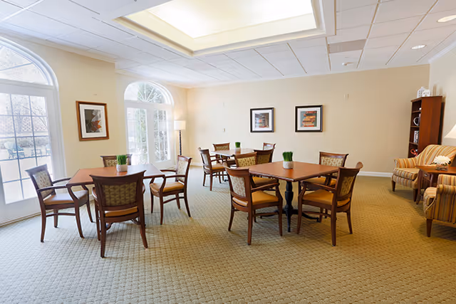A bright and spacious common area with several wooden tables and chairs arranged for seating. The room has large arched windows and glass doors letting in natural light. There are framed pictures on the walls, a tall floor lamp, and a cozy seating area with striped armchairs and a small bookshelf in the corner.