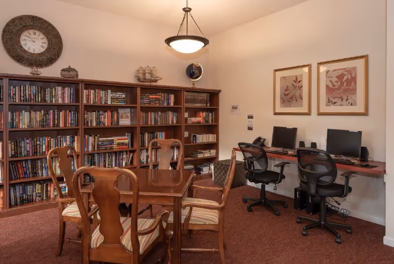 A cozy senior living common area with a wooden table and four chairs in the center. Behind the table is a large bookshelf filled with books, a decorative clock, a model ship, and a globe on top. To the right, there is a desk with two computer workstations and two office chairs. The walls are decorated with two framed pictures featuring leaf designs. The room has warm lighting and carpeted flooring.