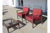 Two red cushioned outdoor chairs and a glass-top coffee table on a concrete patio outside a white building with double doors.