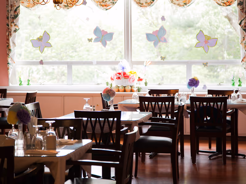 A bright dining room with several wooden tables and chairs arranged neatly. Each table is set with glassware, napkins, and small floral centerpieces. Large windows in the background let in natural light and are decorated with colorful butterfly decals and floral curtains. Outside the windows, greenery is visible.