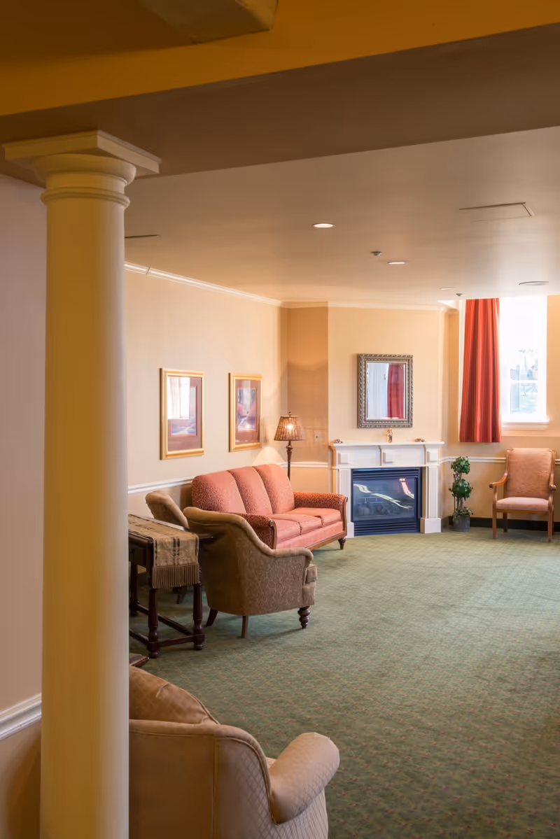 A cozy living room area with a green carpet, beige walls, and a white column in the foreground. The room features a red sofa, two armchairs, a small table with a lamp, a fireplace with a mirror above it, and a window with red curtains letting in natural light.