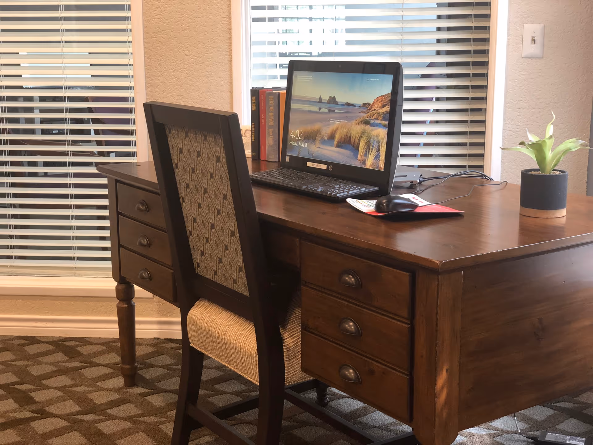 A wooden desk with three drawers on each side and a patterned cushioned chair in front of it. On the desk is a computer monitor displaying a beach scene, a keyboard, a mouse on a red mouse pad, a small potted plant, and several books. The background shows two windows with white blinds partially closed.