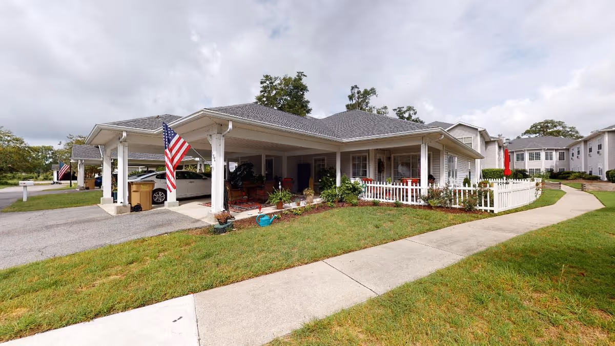 Exterior view of a single-story senior living facility building with a covered carport, American flags, a white picket fence, and a well-maintained lawn with a concrete walkway leading to the entrance. Other similar buildings are visible in the background under a cloudy sky.