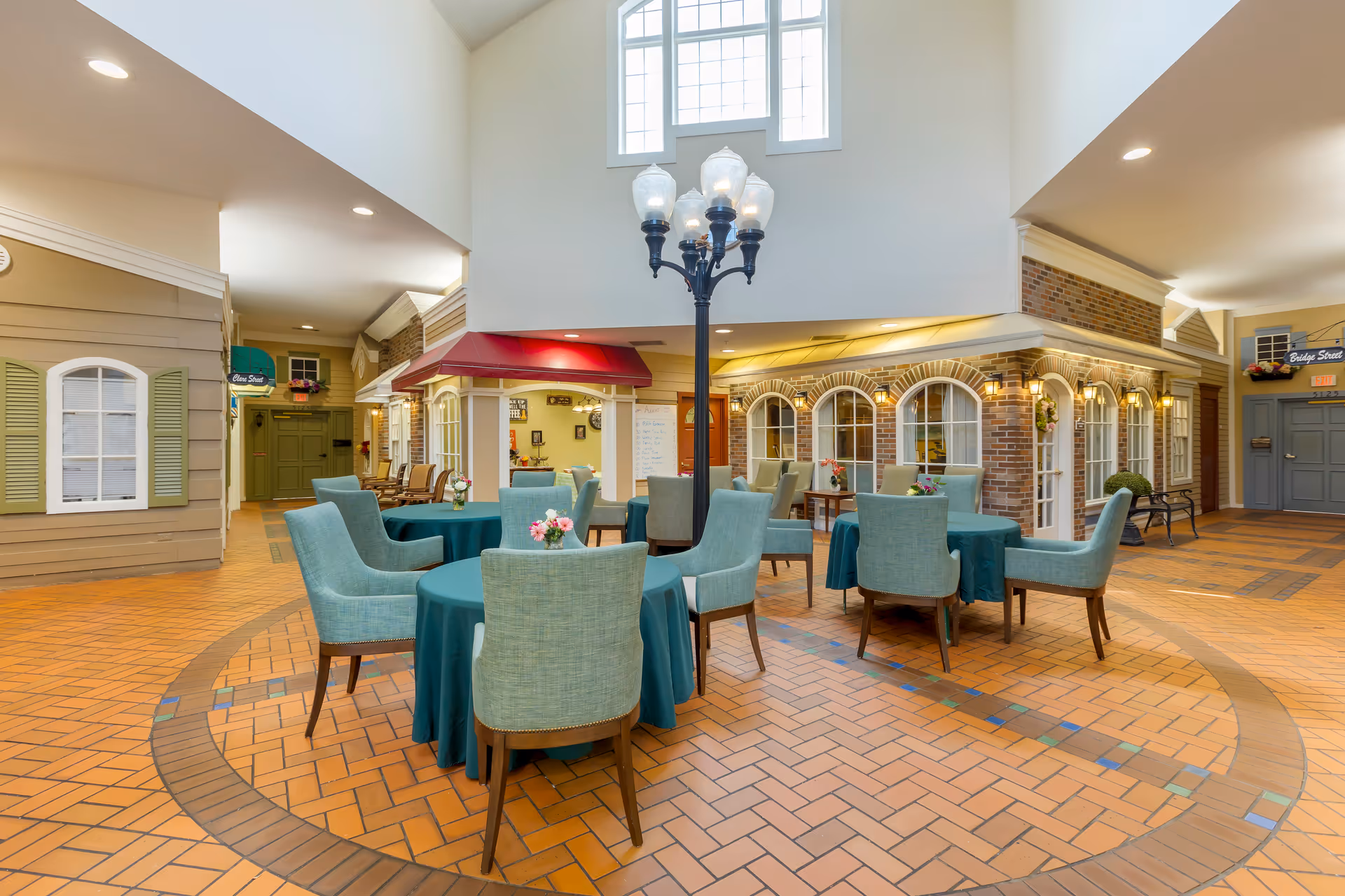 Indoor common area with round tables covered in teal tablecloths and teal upholstered chairs arranged around them. The space features a tall ceiling with large windows, a decorative streetlamp-style light fixture in the center, and building facades with windows and doors resembling a small village street.