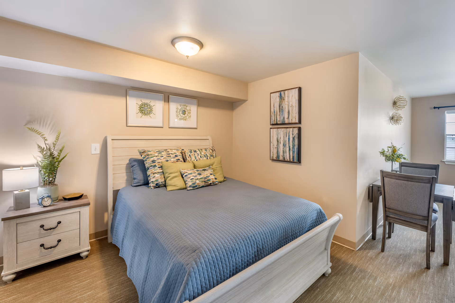 Cozy furnished bedroom with a bed, nightstand, wall art, and a small dining area visible in the background.