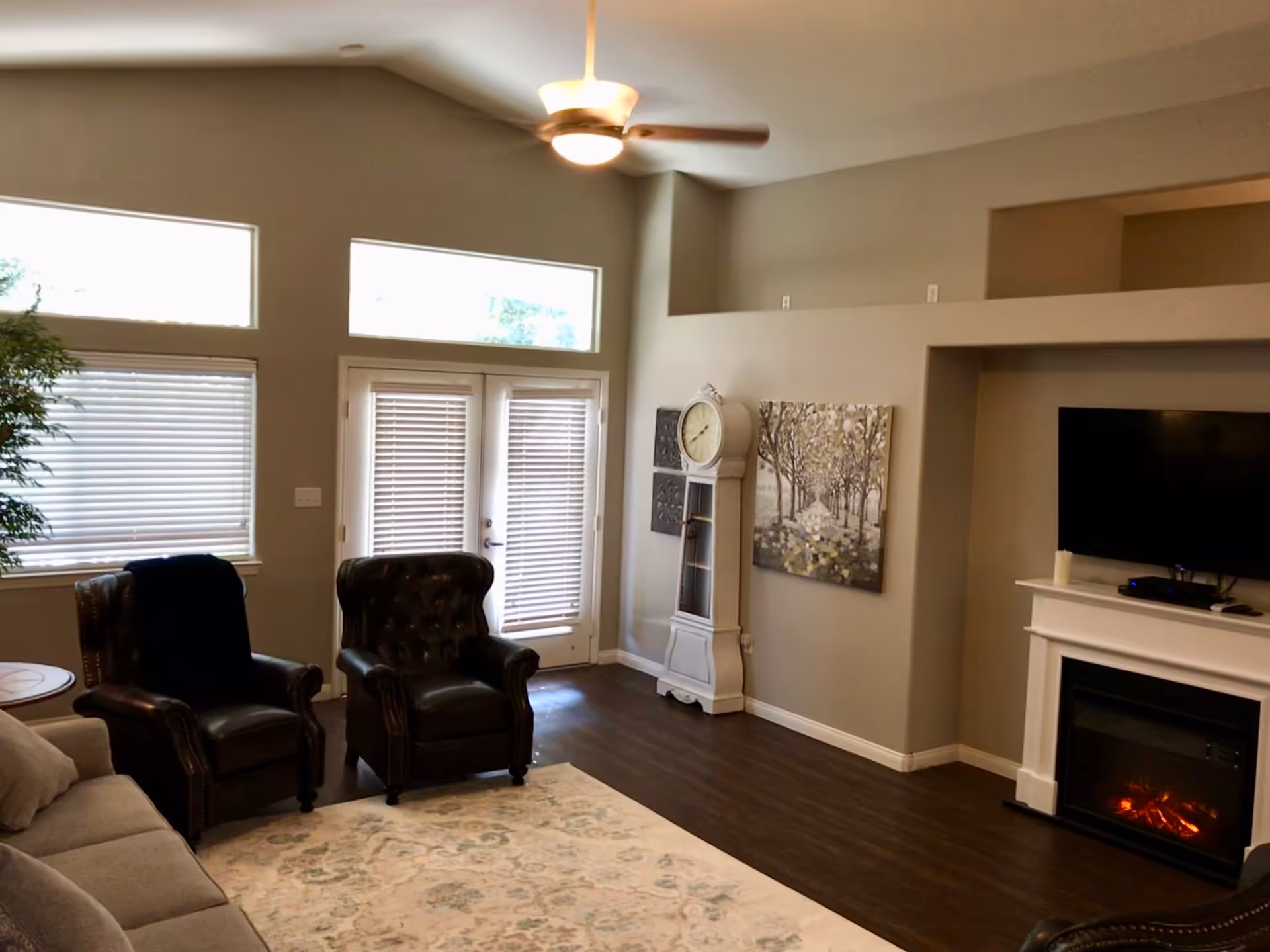 A cozy living room with two dark leather armchairs, a beige sofa, a white grandfather clock, a wall-mounted flat screen TV above a white electric fireplace, and large windows with blinds letting in natural light. The room has wooden flooring and a ceiling fan with a light.