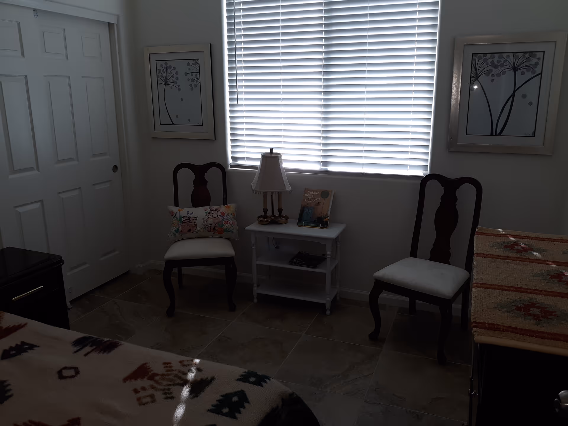 Small bedroom seating area with two wooden chairs flanking a white side table and lamp beneath a window with closed blinds.