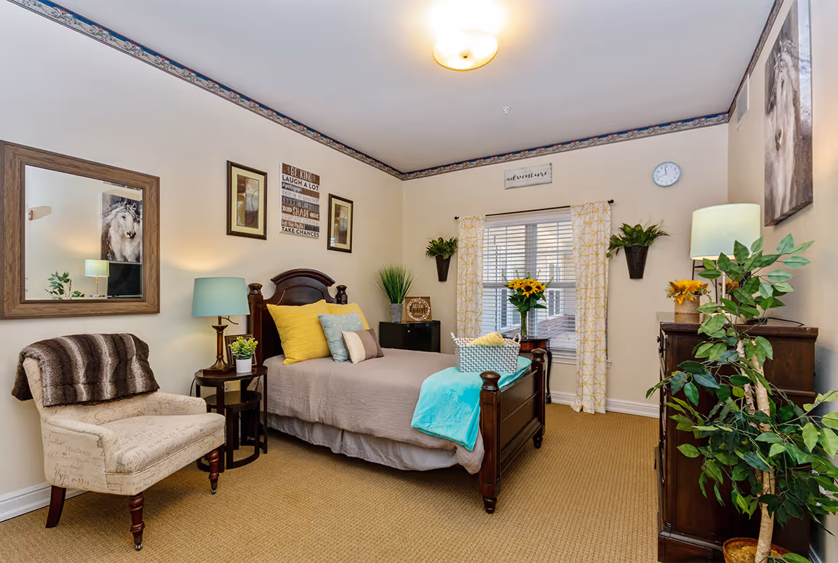 A cozy bedroom with a single wooden bed adorned with yellow and blue pillows and a light brown bedspread. Next to the bed is a small round table with a lamp and a potted plant. There is a beige upholstered chair with a brown throw blanket draped over it. The room features a large window with yellow patterned curtains, two wall-mounted plants, and framed artwork on the walls. A wooden dresser with a lamp and a potted plant is on the right side, and a large leafy plant is in the corner. The carpet is beige, and the walls are light-colored with a decorative border near the ceiling.