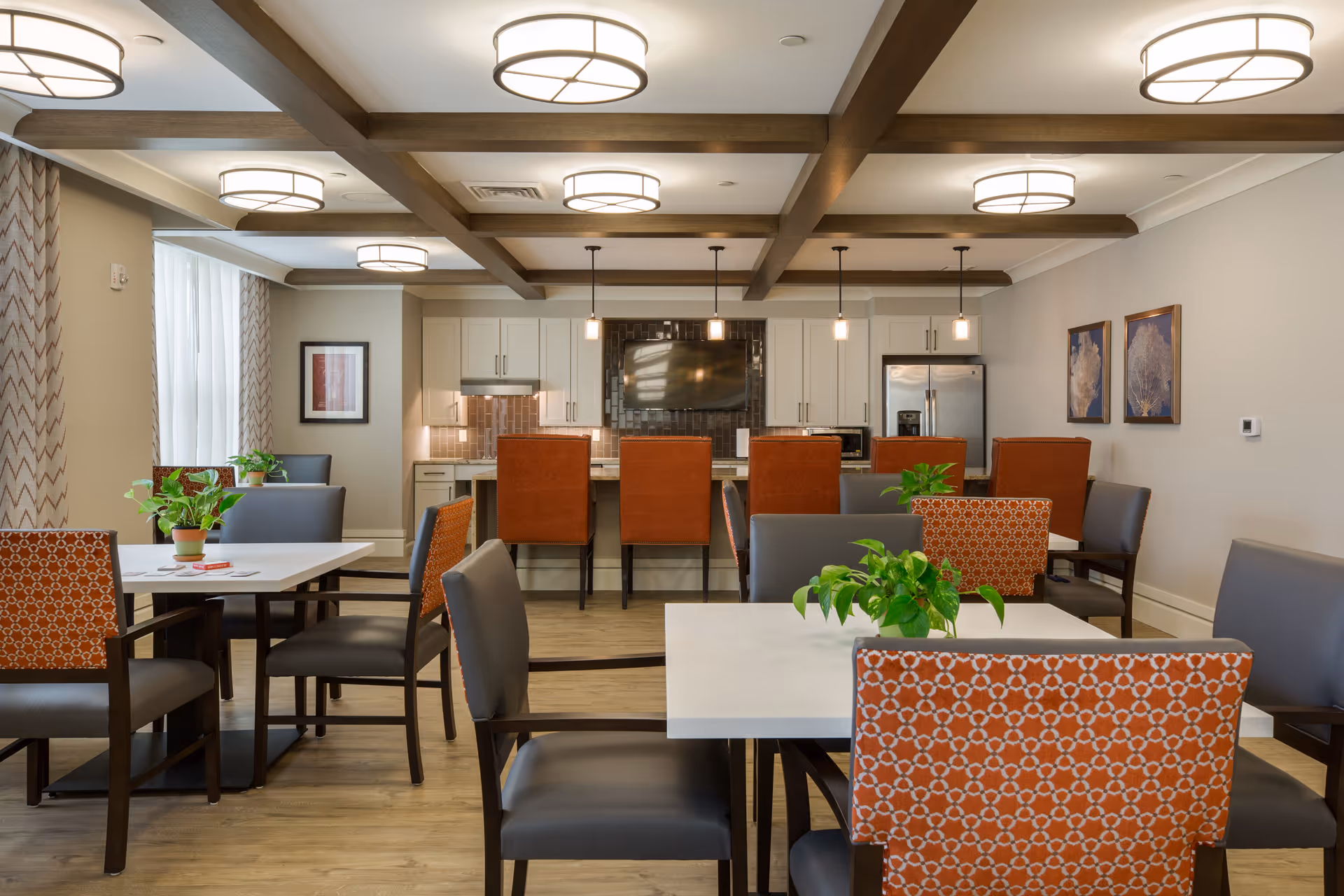 Communal dining room with multiple tables and patterned chairs, potted plants, and a kitchen island with barstools beneath exposed ceiling beams and pendant lights.