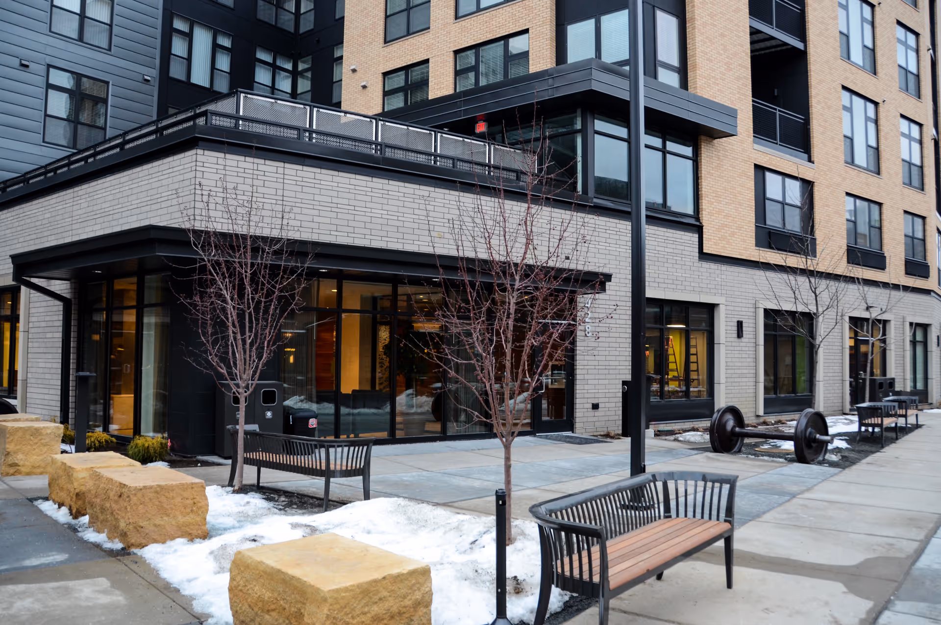 Exterior front entrance of a modern brick-and-glass senior living building with benches, small trees, and patches of snow.