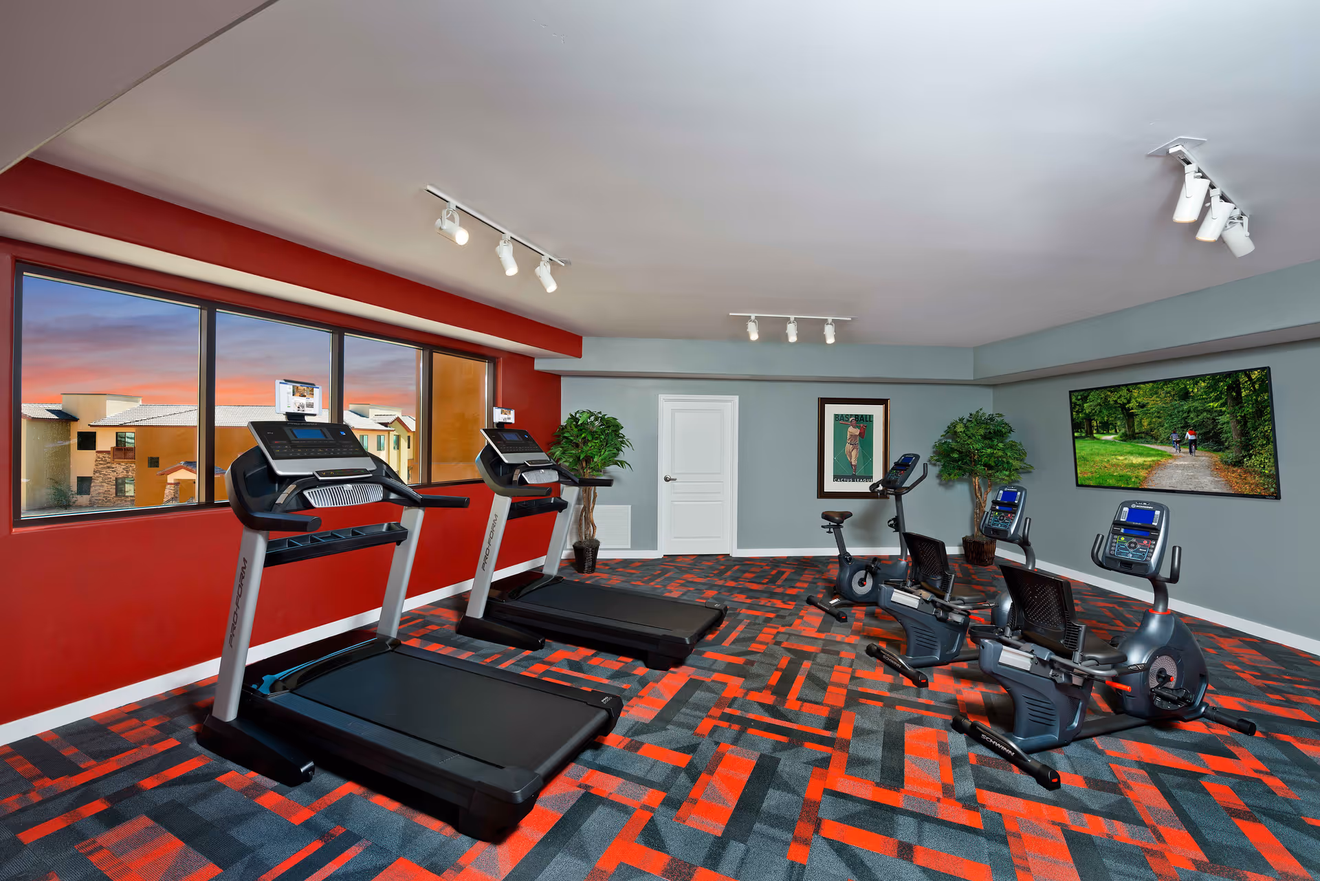 Exercise room with treadmills and recumbent bikes, patterned red-and-gray carpet, large windows and a wall-mounted TV.