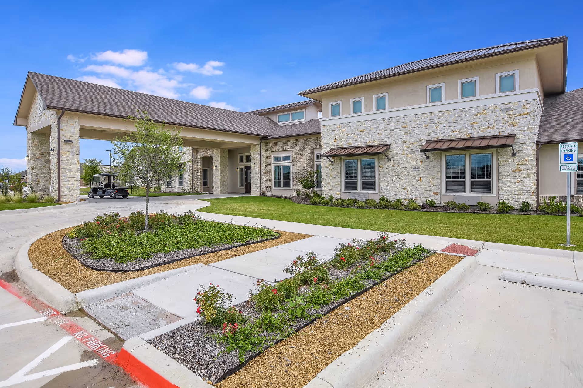 Exterior view of a senior living facility building with stone walls and a covered entrance. There is a landscaped area with small bushes and flowers in front, a concrete driveway, and a reserved parking sign for handicapped parking. The sky is clear with a few clouds.