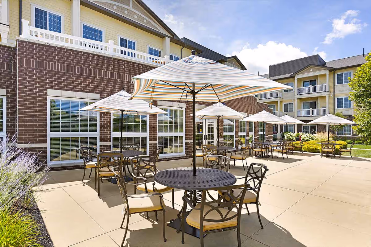 Outdoor patio area at StoryPoint Pickerington with multiple round metal tables and chairs, each table shaded by a large striped umbrella. The patio is adjacent to a brick and yellow siding building with large windows. The sky is blue with some clouds.