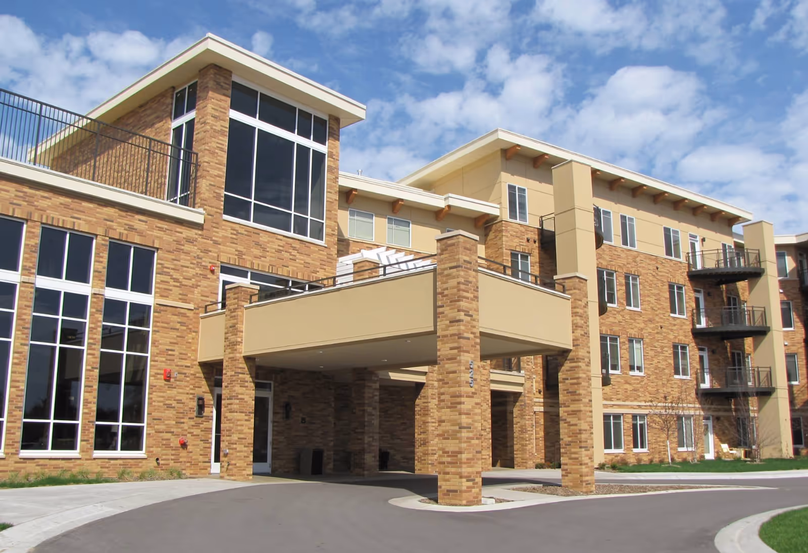 Exterior view of a multi-story senior living facility building with brick and beige siding, large windows, balconies, and a covered entrance driveway under a partly cloudy sky.