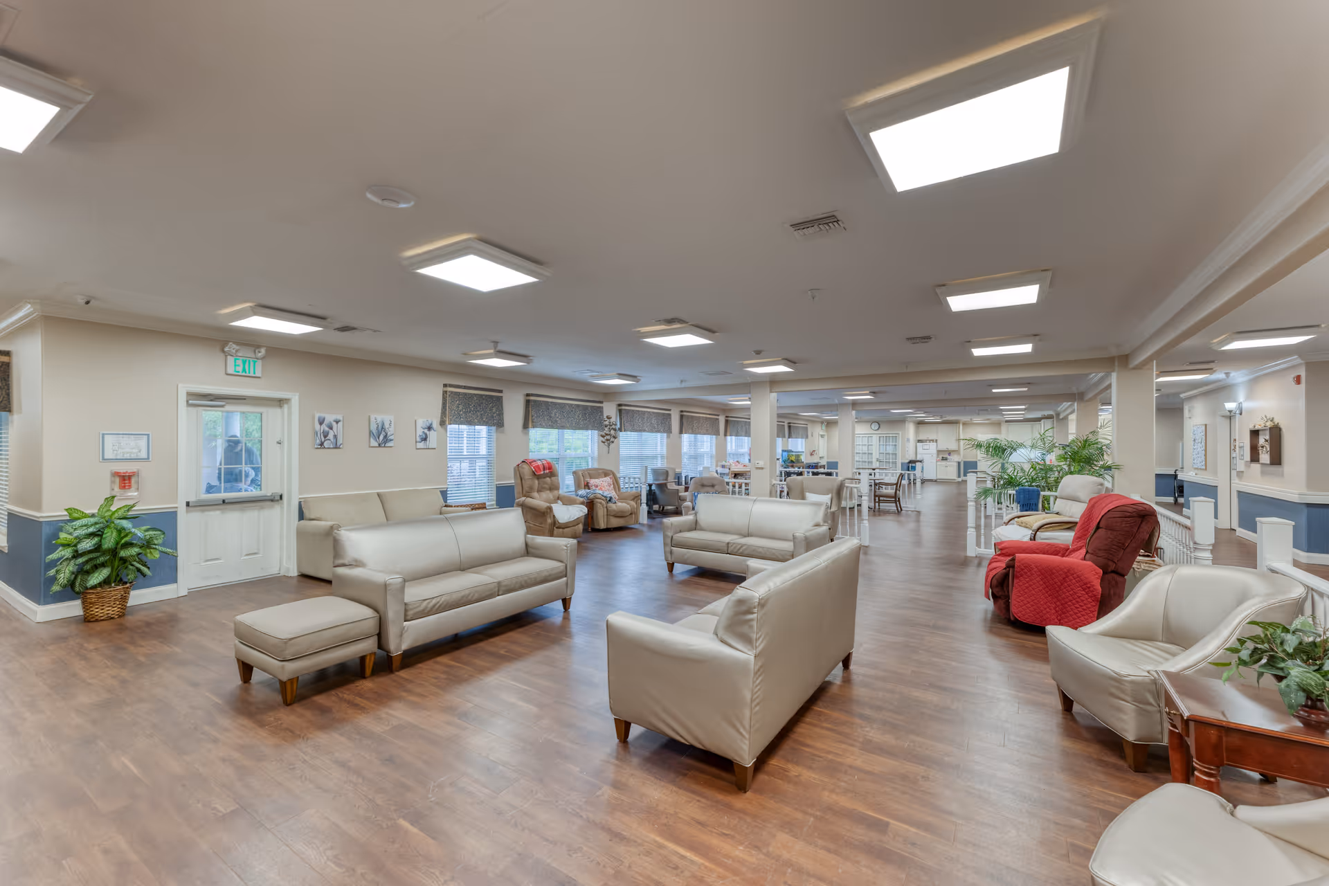 A spacious, well-lit common area in a senior living facility featuring multiple beige leather sofas and armchairs arranged on a wooden floor. The room has large windows with blinds, several potted plants, and overhead square ceiling lights. There are also recliner chairs with red blankets and a door with an exit sign above it on the left side.