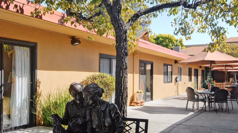Outdoor courtyard of a nursing facility with a bronze statue of an elderly couple on a bench, a tree, and patio tables with umbrellas beside the building.