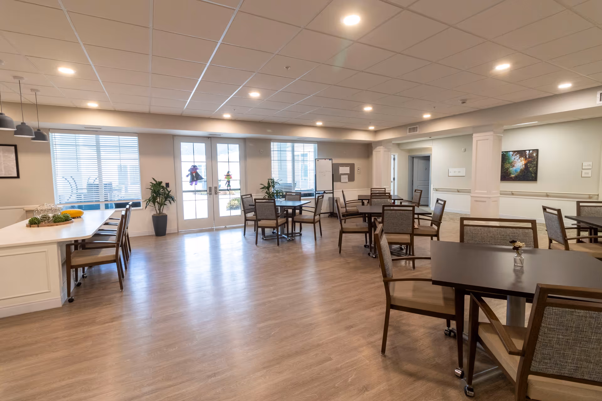 A spacious, well-lit dining area with multiple tables and chairs arranged neatly. There are large windows and glass doors letting in natural light, with some decorative plants near the windows. The floor is wooden, and the ceiling has recessed lighting. A counter with chairs is visible on the left side, and there is a whiteboard and framed artwork on the walls.