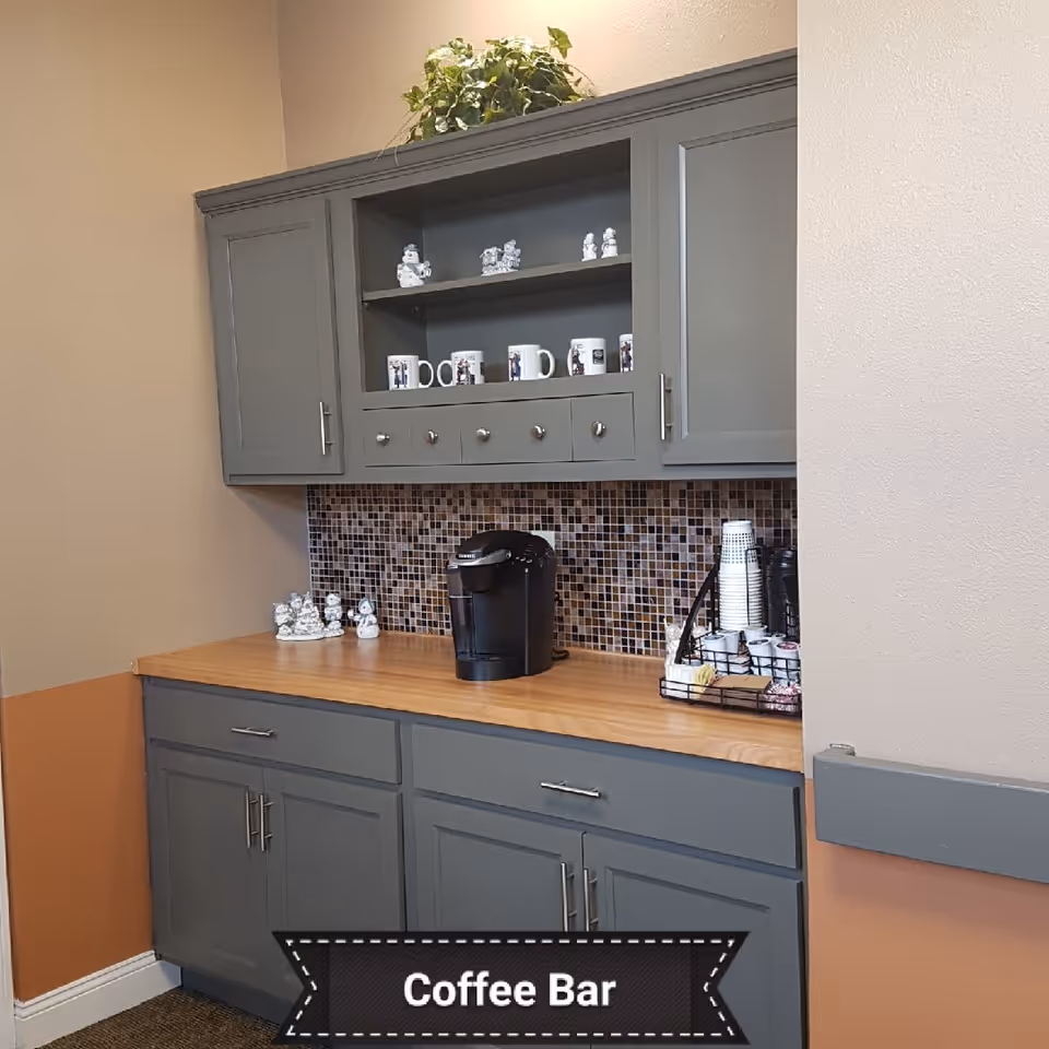 A coffee bar area with gray cabinets and a wooden countertop. There is a coffee maker, a tray with disposable cups, creamers, and sugar packets. The backsplash is made of small square mosaic tiles in various shades of brown. Decorative items and mugs are displayed on the upper shelves, and a green plant is on top of the cabinet.