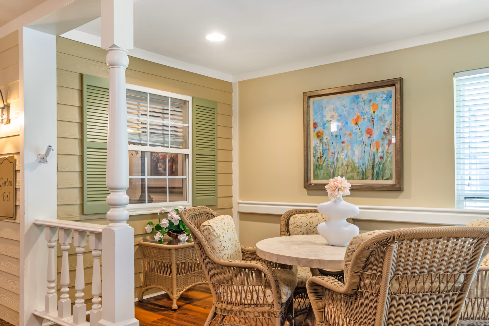 Cozy indoor seating area with a round table and wicker chairs, a decorative vase, a floral painting on the wall, and a window with green shutters.