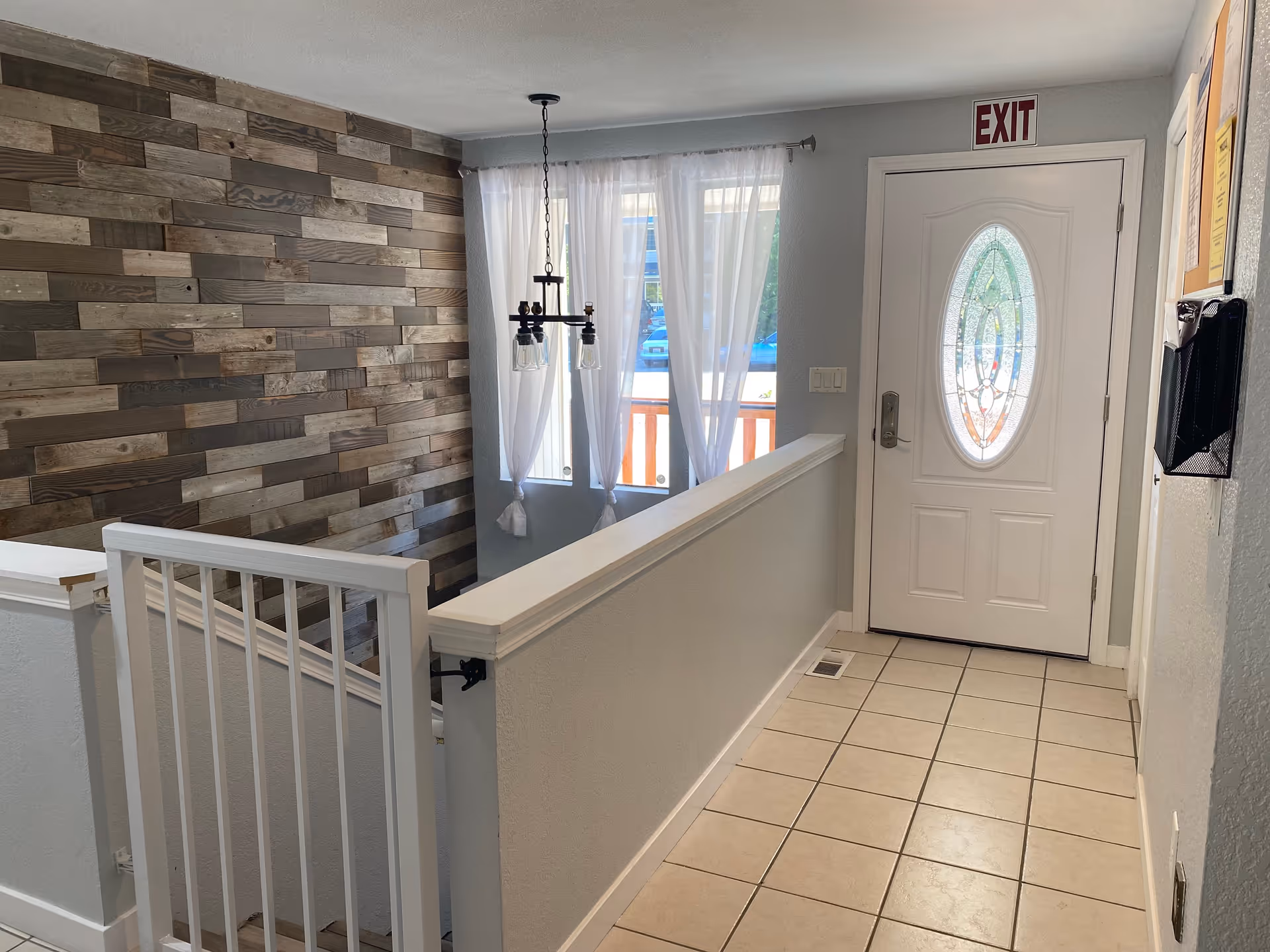 Interior hallway with tiled floor, a white door with decorative glass, an exit sign above the door, a wooden accent wall, a hanging light fixture, and a stairway with a white railing.