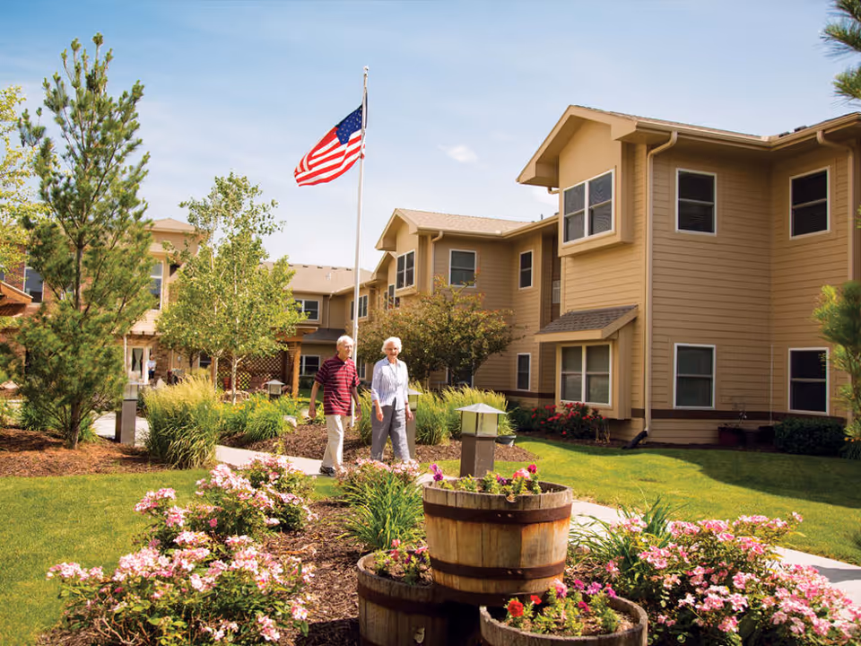 Two elderly people walking on a paved path through a landscaped garden area with flowers and greenery, in front of a beige multi-story residential building under a clear blue sky with an American flag on a flagpole.