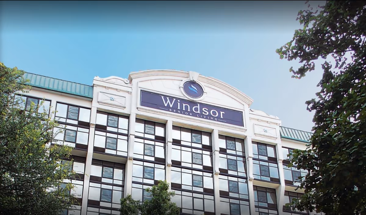 Exterior view of a multi-story senior living facility building with a sign that reads 'Windsor Senior Living' at the top center, surrounded by trees under a clear blue sky.