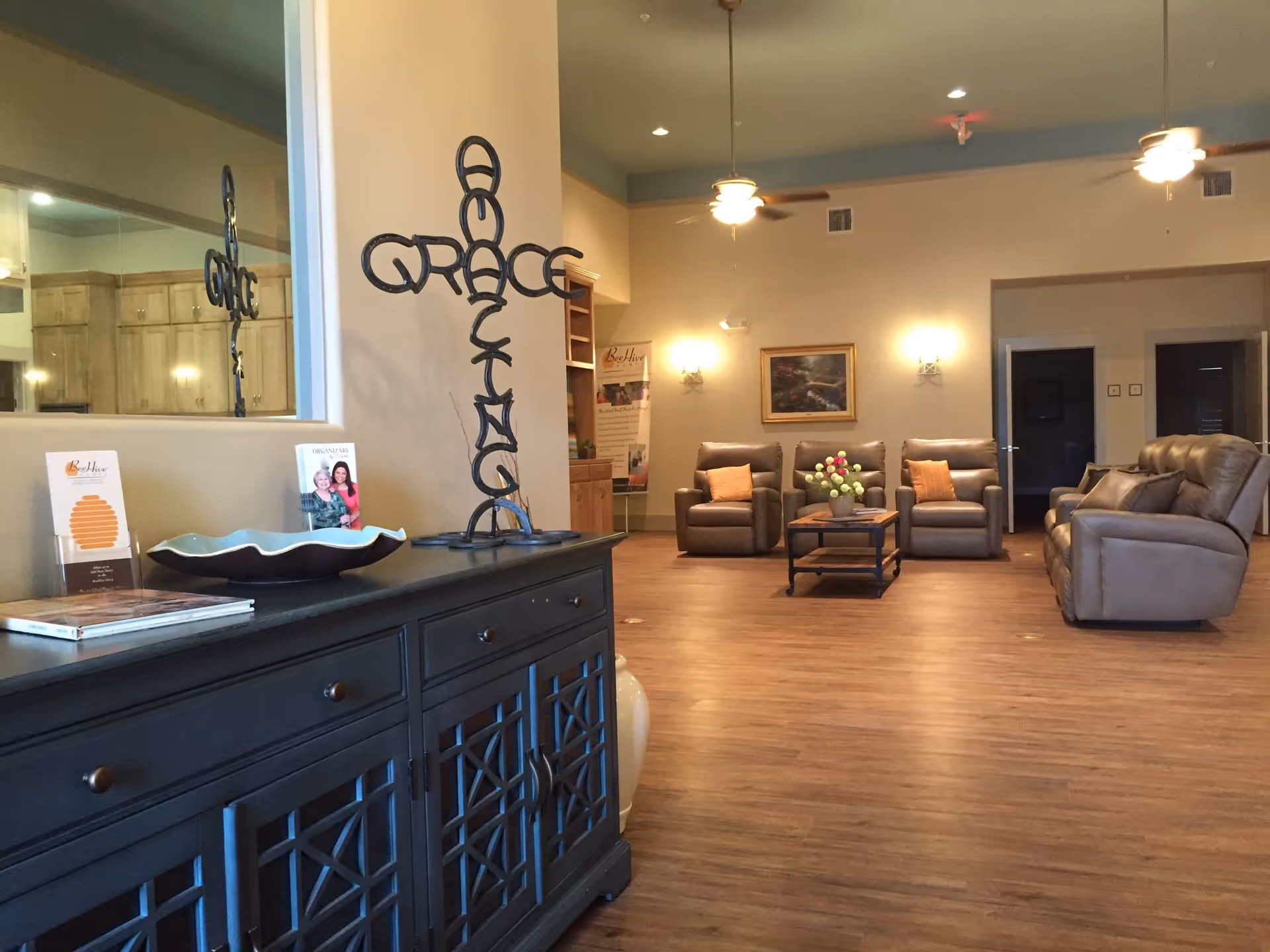 A spacious living room area with wooden flooring, beige walls, and ceiling fans. There are three brown leather armchairs with orange cushions and a matching sofa. A coffee table with a flower arrangement is in the center. On the left side, there is a dark wooden cabinet with decorative items including a metal cross sculpture spelling the word 'GRACE' and brochures. The room is softly lit with wall sconces and ceiling lights.