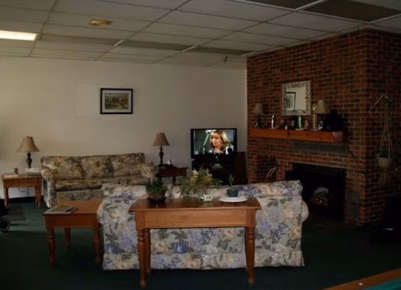 A cozy living room with floral patterned sofas and wooden tables. There are two table lamps on side tables, a TV showing a woman, and a brick fireplace with a wooden mantel decorated with lamps and a mirror. The room has a green carpet and a framed picture on the wall.