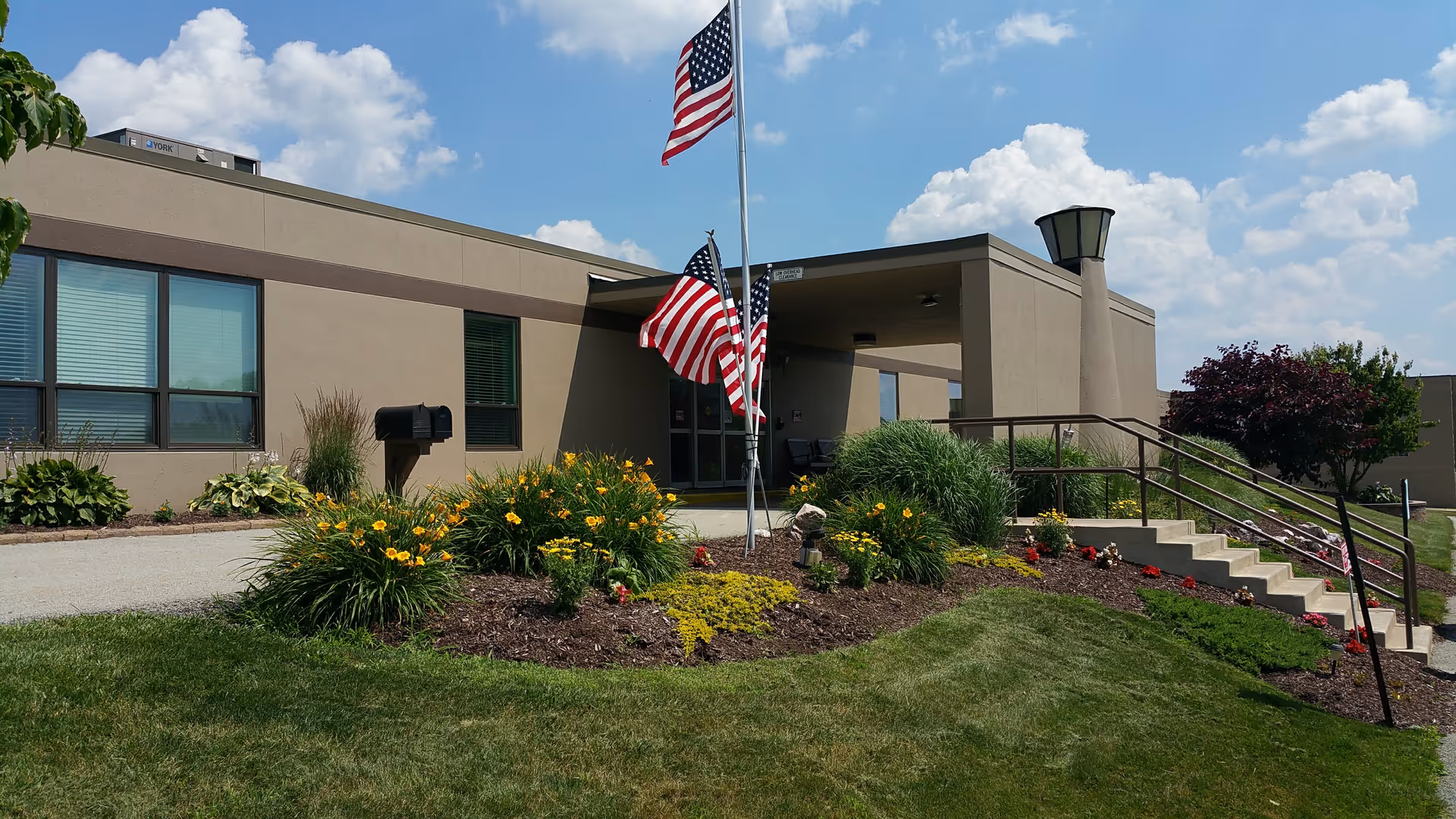 Exterior view of a single-story building with beige walls and several windows. In front of the building is a landscaped garden with green shrubs, yellow flowers, and a small American flag display on a flagpole. There are stairs with a handrail leading up to the entrance, and the sky is partly cloudy with blue patches.