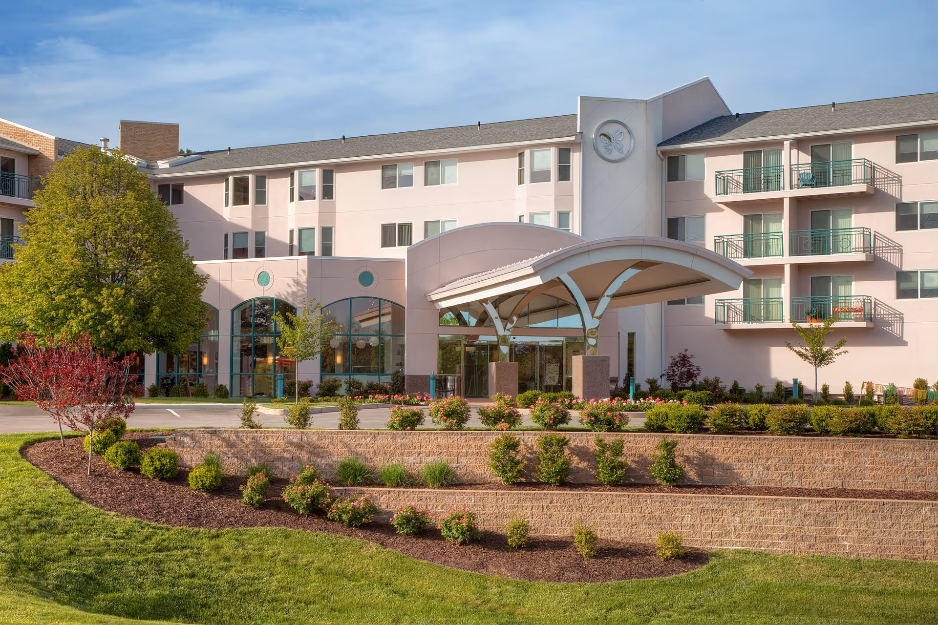 Exterior view of Chesterfield Villas, a multi-story senior living facility with balconies, large windows, and a covered entrance. The building is surrounded by landscaped gardens with green grass, shrubs, and trees under a partly cloudy sky.