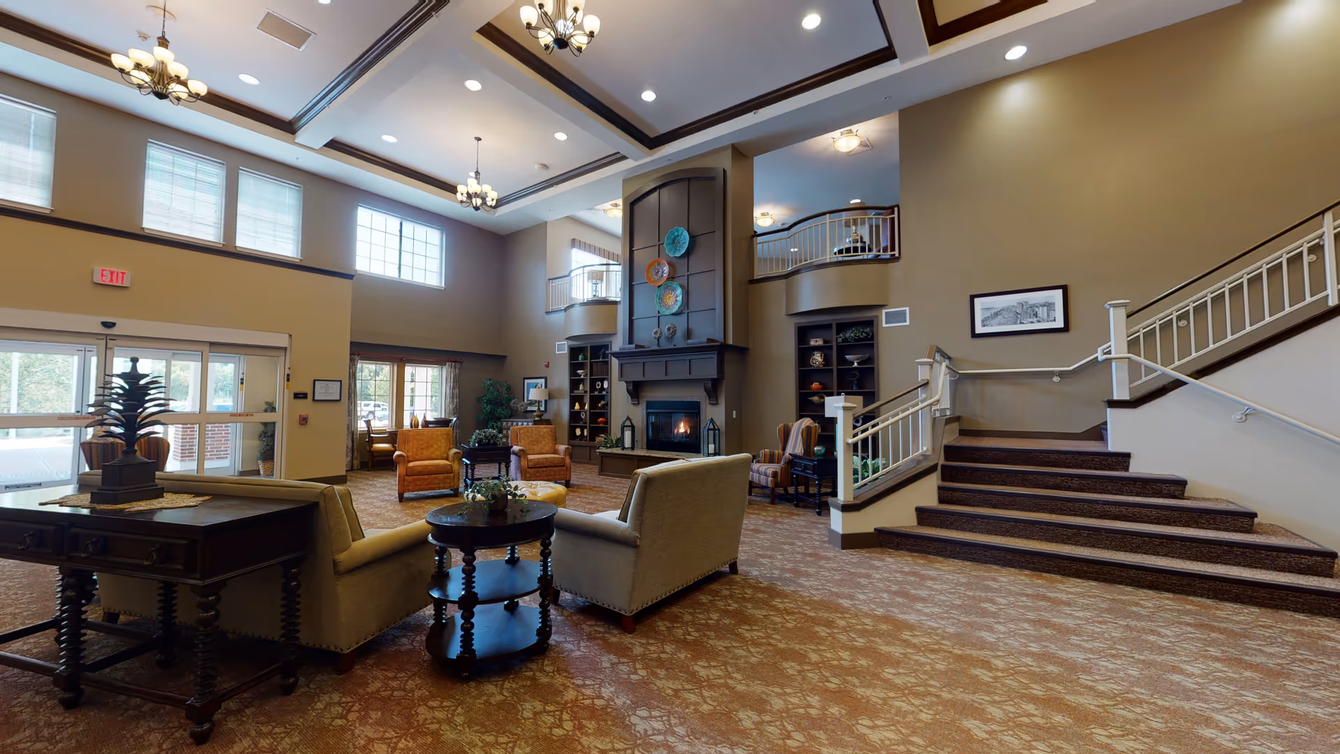 Spacious and well-lit common area in a senior living facility with high ceilings, large windows, and multiple seating arrangements including sofas and armchairs around a central fireplace. There is a staircase leading to an upper level with a balcony overlooking the room. The decor includes chandeliers, framed artwork, and decorative plates on the fireplace mantel.