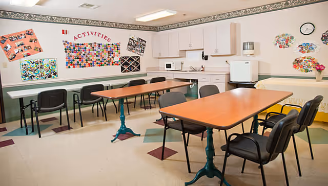 A bright activity room with tables and chairs arranged for group activities. The room features a kitchenette area with cabinets, a microwave, and a small refrigerator. The walls are decorated with colorful bulletin boards labeled 'ACTIVITIES' and various artwork and photos. The floor has a patterned linoleum design.