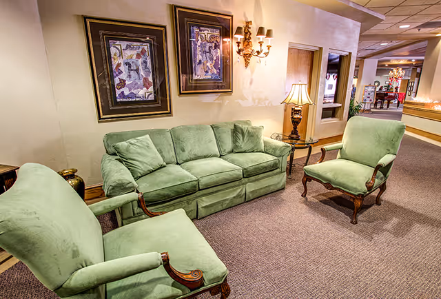A cozy seating area in a senior living facility featuring a green upholstered sofa and two matching armchairs arranged on a carpeted floor. The wall behind the sofa has two framed abstract paintings and a decorative wall sconce with two lights. A table lamp on a side table is positioned next to the sofa, and the hallway in the background shows more seating and decor.