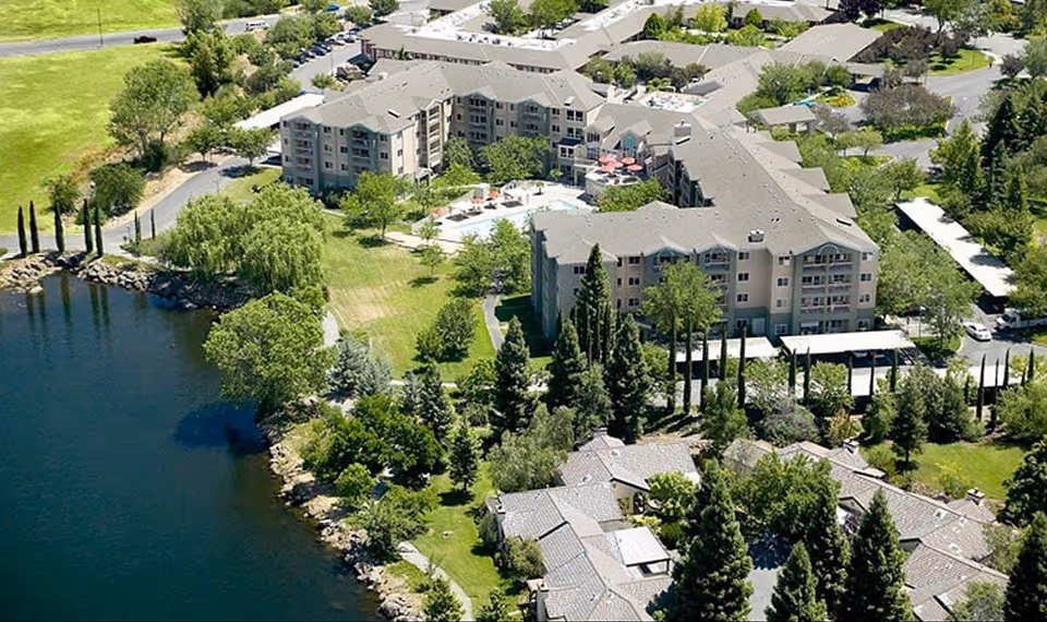 Aerial view of Westmont of Chico senior living facility showing multiple buildings surrounded by trees and greenery, with a large pond on the left side and pathways connecting the buildings.