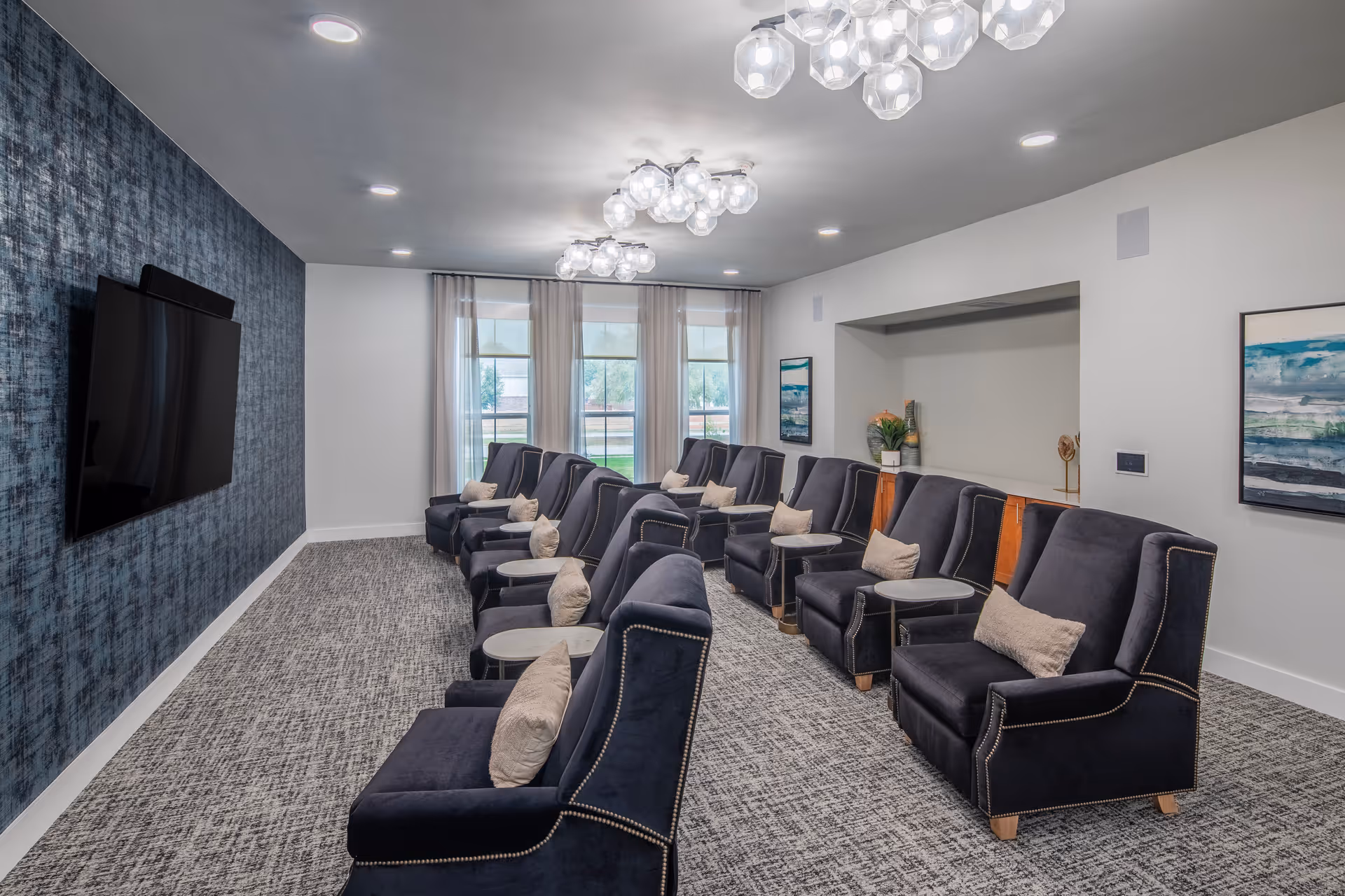 A modern lounge area with two rows of dark blue armchairs, each with a small side table and a beige pillow. The room has a large flat-screen TV mounted on a textured blue accent wall, three large windows with sheer curtains, and contemporary ceiling light fixtures. The carpet is patterned in gray tones, and there are framed artworks on the walls.